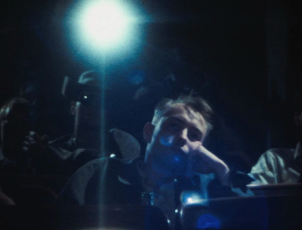 Nekromantik (1988) movie still: medium — A dimly lit room filled with people sitting in dark rows of seats. A young man rests his chin on his hand…