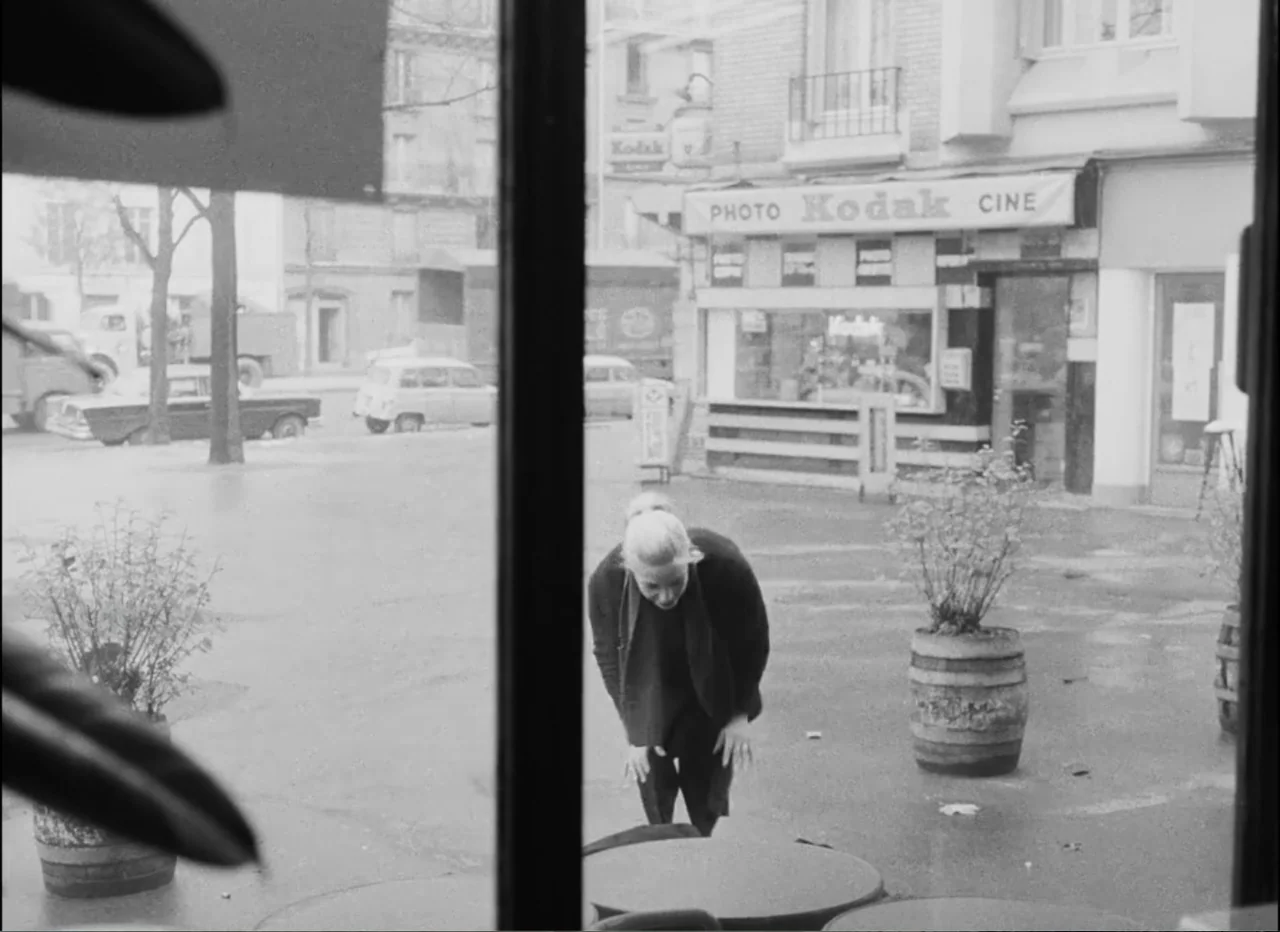 Masculin Féminin (1966) movie still: medium — A woman leans over a round table on a sidewalk outside a retail shop. Cars and buildings are visible…