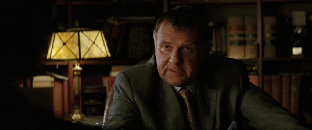 Batman Begins (2005) movie still: medium — A dimly lit room features bookshelves filled with volumes in the background. An older man sits at a desk…