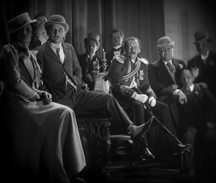 The Threepenny Opera (1931) movie still: medium — A dimly lit meeting space features dark wooden furniture and rows of seated individuals. A man in…