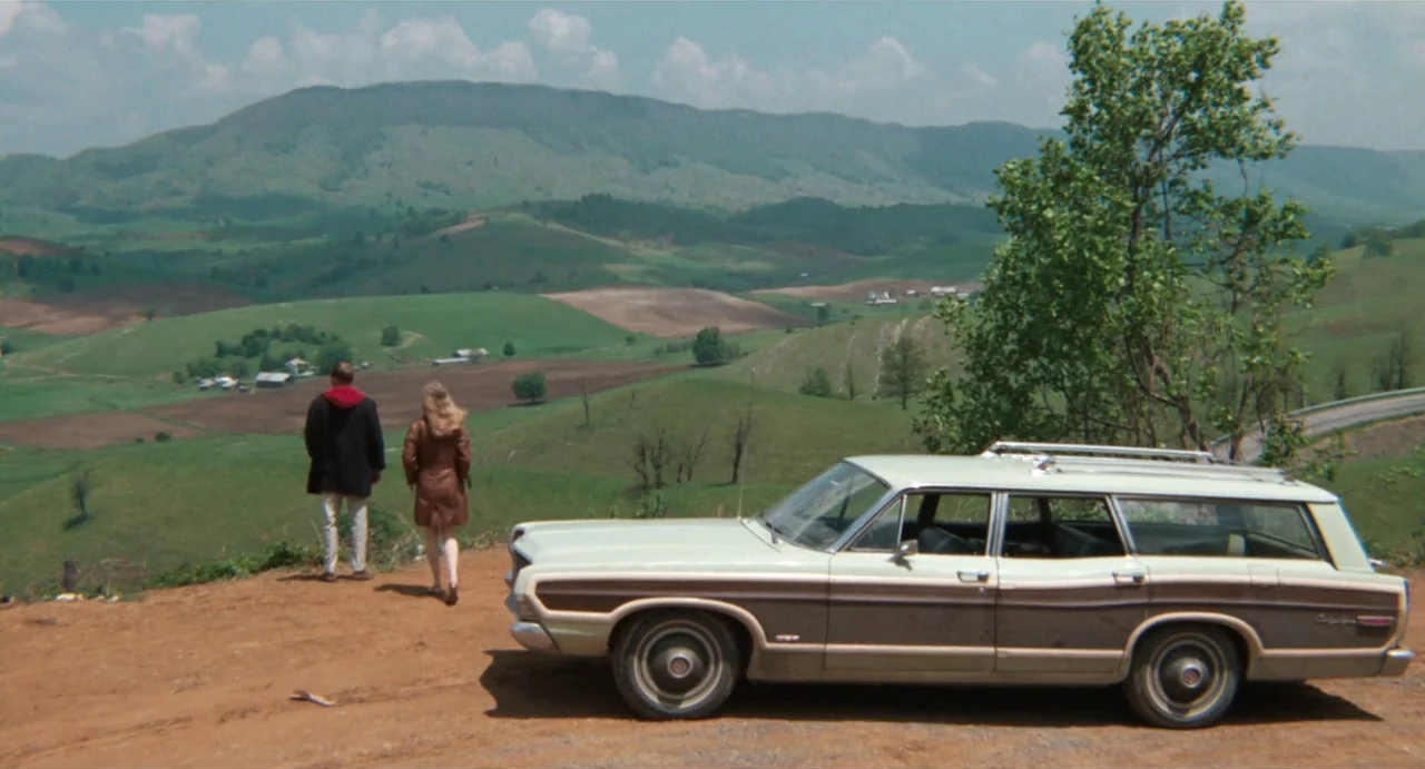 The Rain People (1969) movie still: wide — A dirt overlook faces a wide expanse of rolling green hills under a cloudy sky. A man and a woman stand…