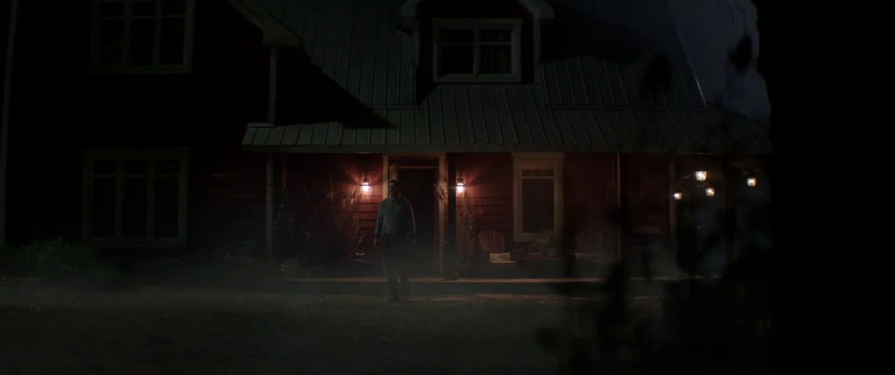 Pet Sematary (2019) movie still: wide — A dark wooden house at night features a porch illuminated by two glowing wall lamps. A man stands motionless…