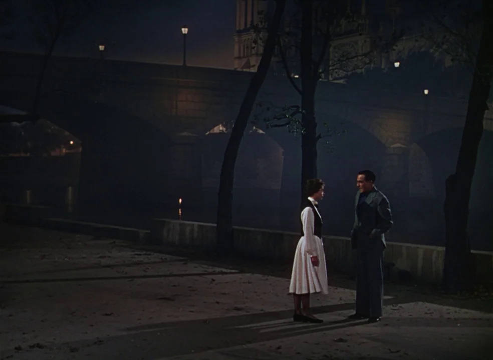An American In Paris (1951) movie still: wide — A night view of a river embankment with a large stone bridge in background. A man and woman stand…