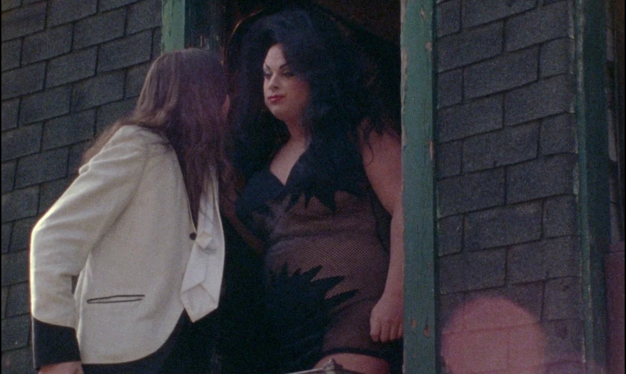 Female Trouble (1974) movie still: medium — A fire escape doorway features a dark brick wall backdrop. A person in a blazer speaks to a drag…