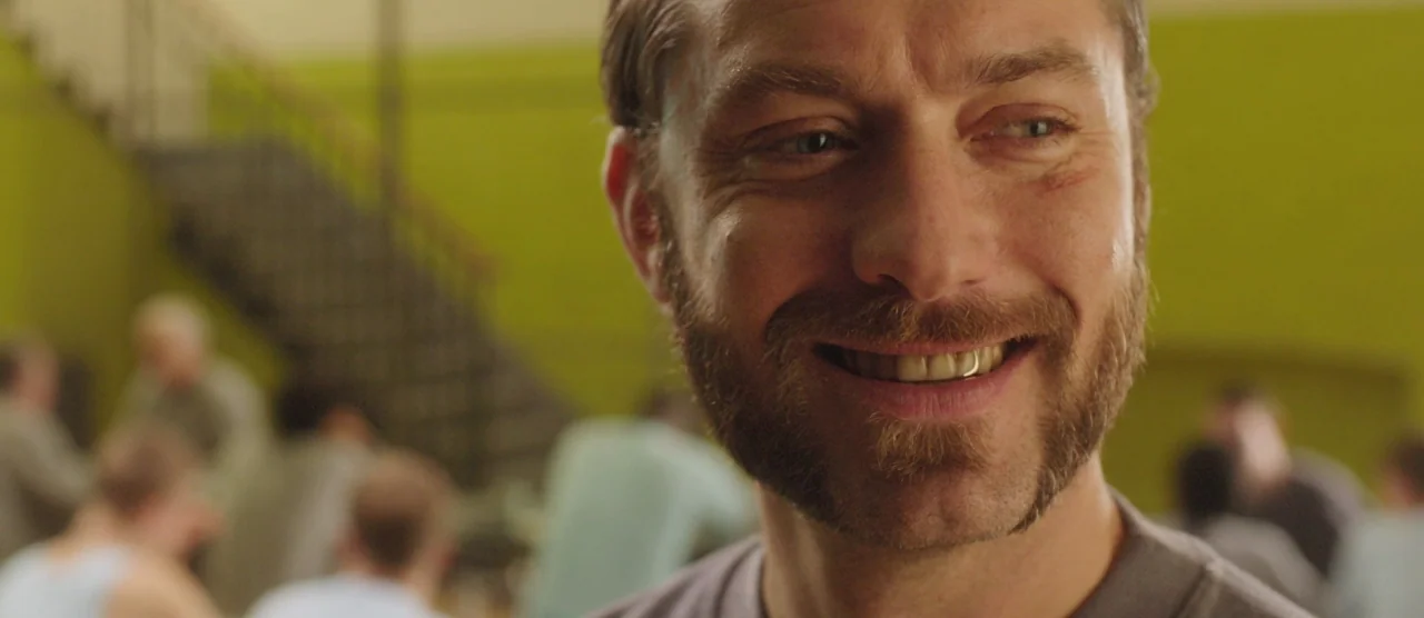 Dom Hemingway (2013) movie still: closeup — A cafeteria interior features a lime-green wall and a blurry staircase in the background. A man smiles…