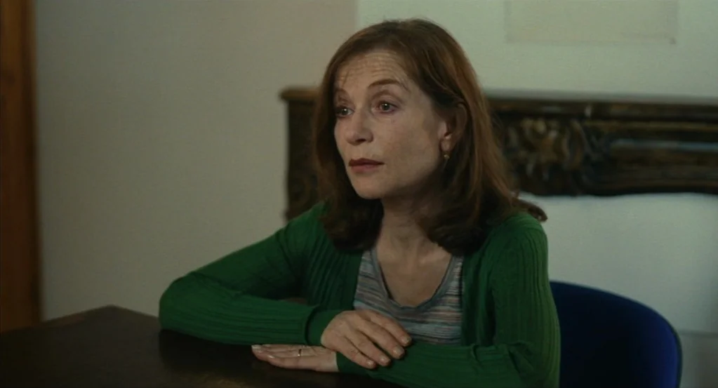 Things to Come (2016) movie still: medium — A woman sits at a wooden table in a room with a decorative fireplace mantle. She rests her hands on the…