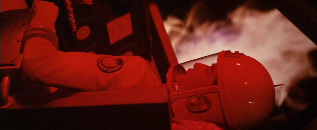 Destroy All Monsters (1968) movie still: medium — An astronaut in a white suit sits in a darkened cockpit under red light. The pilot wears a rounded…
