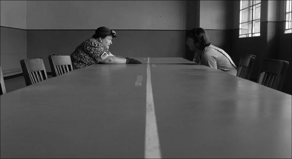 The Man Who Wasn’t There (2001) movie still: wide — A spartan room features a very long table separating two seated women in profile. Tall, barred…