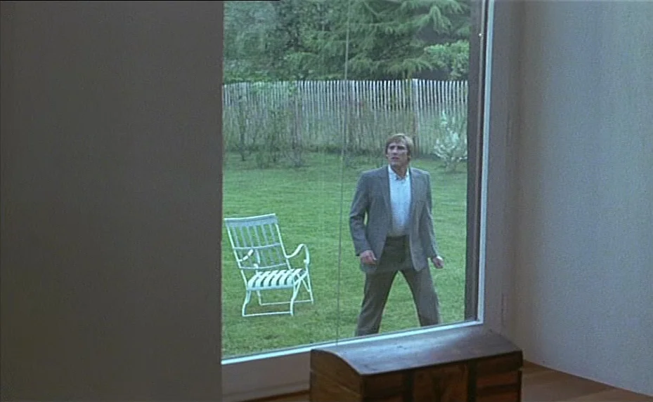 The Woman Next Door (1981) movie still: medium — A modern minimalist interior connects to a green lawn via a large glass window. A man stands on the…