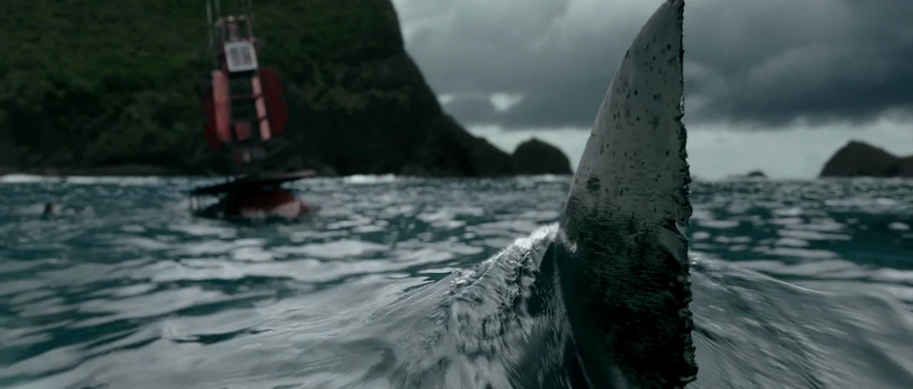 The Shallows (2016) movie still: closeup — A shark dorsal fin cuts through choppy grey water near a red navigation buoy. Rocky cliffs and a dark,…