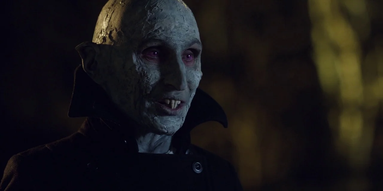 Jakob’s Wife (2021) movie still: closeup — A dark outdoor setting with blurred silhouettes of trees in the background. A pale creature with pointed…