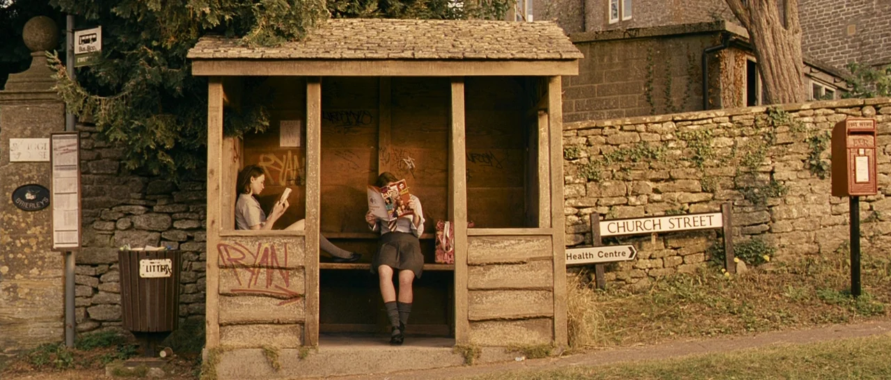 Tamara Drewe (2010) movie still: fullbody — A wooden bus shelter with graffiti sits along a stone wall on a rural street. Two teenage girls in…