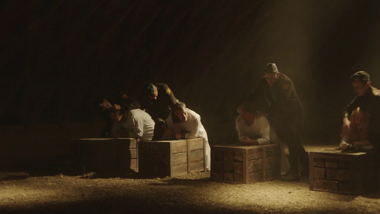 Infinity Pool (2023) movie still: wide — A dimly lit, vast attic space features wooden rafters overhead and a dirt floor. Several soldiers in…