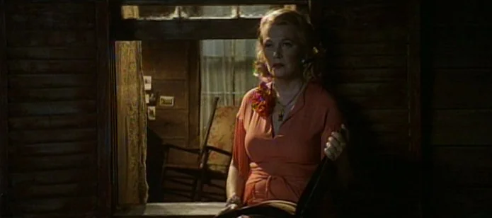The Neon Bible (1995) movie still: medium — A woman sits on a porch at night in front of a dark window. She holds an open book while looking…