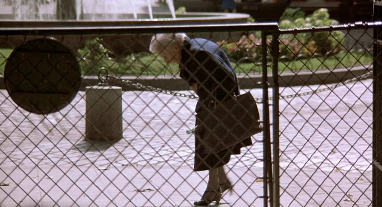 Three Colours: Blue (1993) movie still: fullbody — A park pathway is enclosed by a prominent chain-link fence with a fountain behind. An elderly…