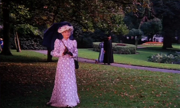 Katie Tippel (1975) movie still: fullbody — A well-maintained public park features mature trees with autumn leaves on the grass. A woman in an…