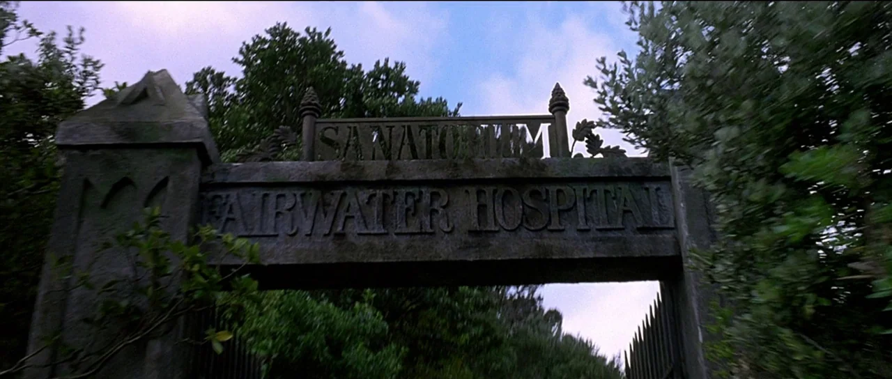 The Frighteners (1996) movie still: establishing — A stone archway marked Fairwater Hospital Sanatorium stands amidst thick, dark green foliage. The…