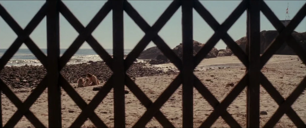 Inception (2010) movie still: wide — A wooden lattice fence frame separates the foreground from a wide, sandy beach. Rocks line the shoreline where…
