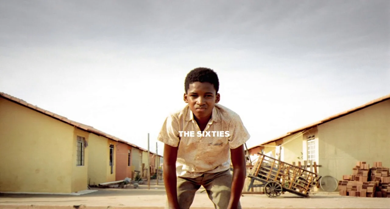 City of God (2002) movie still: medium — Two rows of simple stucco houses line a dusty road under a bright sky. A young boy stands centered in the…