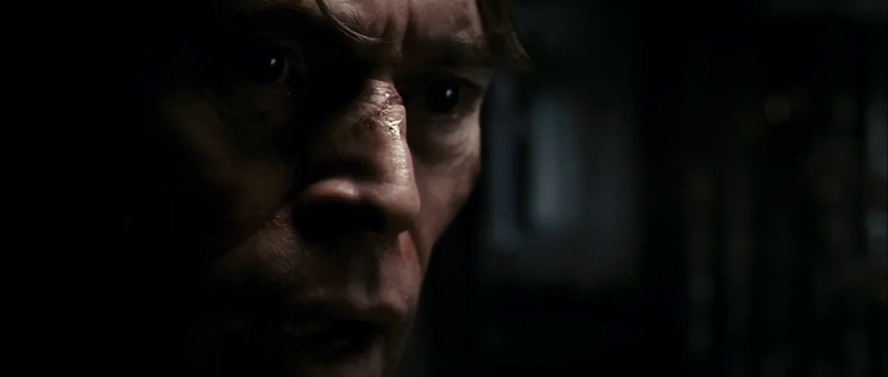 Antichrist (2009) movie still: closeup — A man's face emerges from the darkness in a dimly lit interior space. He gazes off-camera with his scarred…
