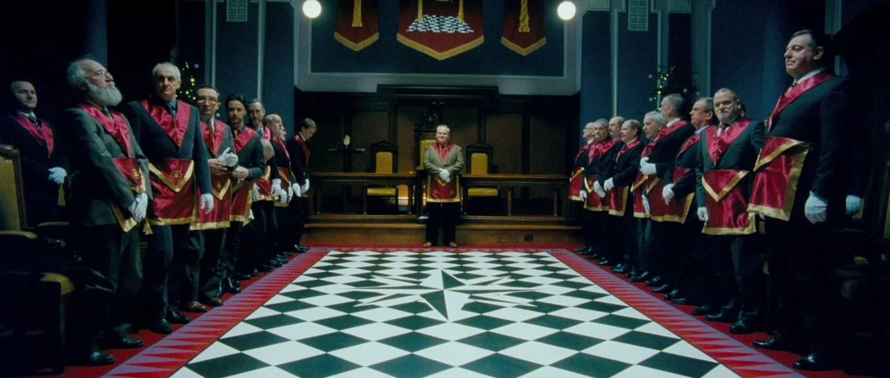 Filth (2013) movie still: wide — A formal meeting hall features a black and white checkerboard floor and blue walls. A group of men in suits and…