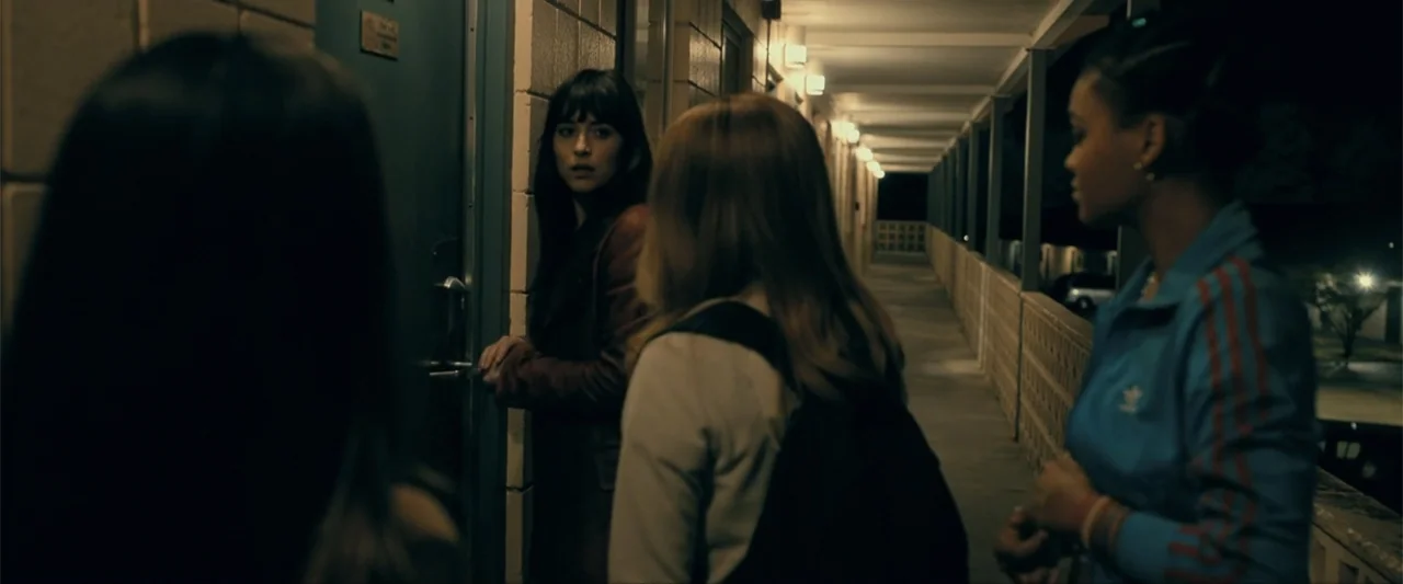 Madame Web (2024) movie still: medium — A night view of an exterior motel walkway with repeating illuminated wall lights. Three women stand near a…