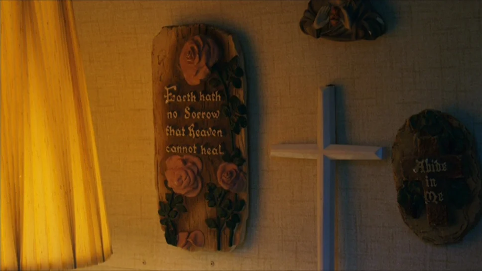 Youth In Revolt (2009) movie still: closeup — A wall decorated with religious plaques and a wooden cross is shown close up. A warm lampshade sits on…