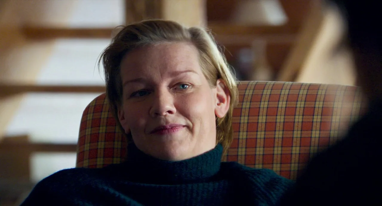Anatomy of a Fall (2023) movie still: closeup — A person sits in a patterned fabric chair inside a brightly lit room. A woman with short blonde hair…