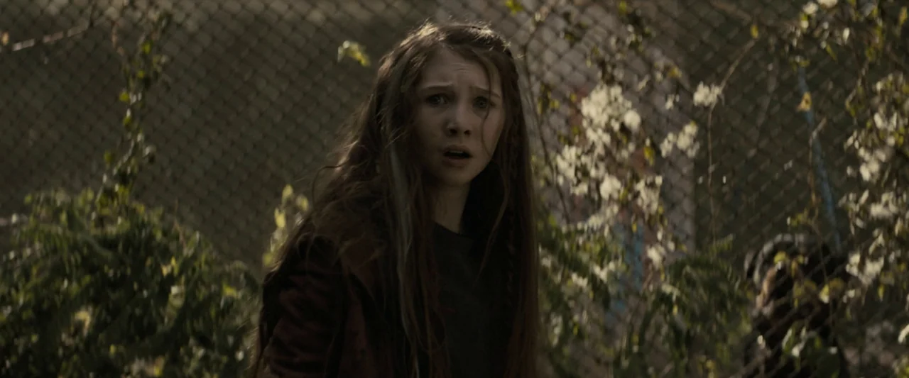 Don’t Breathe 2 (2021) movie still: medium — A young girl stands in front of a chain-link fence overgrown with leafy green vines. She looks…