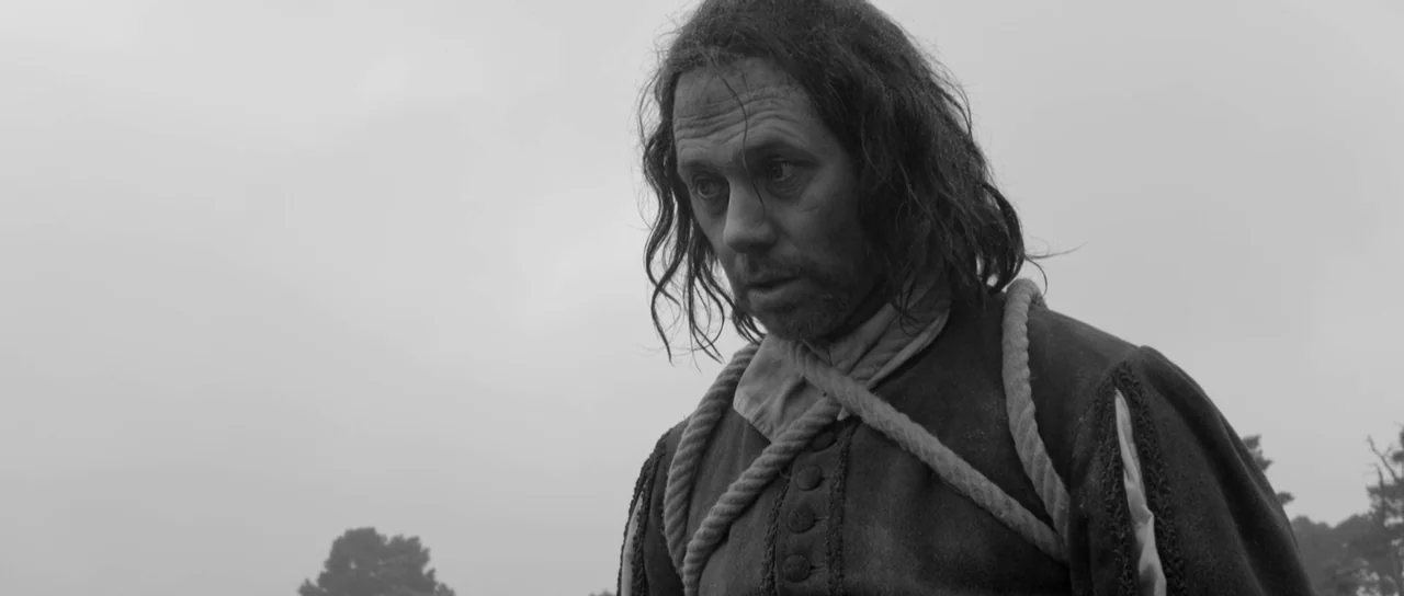 A Field In England (2013) movie still: medium — An open field under a flat grey sky. A man with a rope around his neck stares downward.