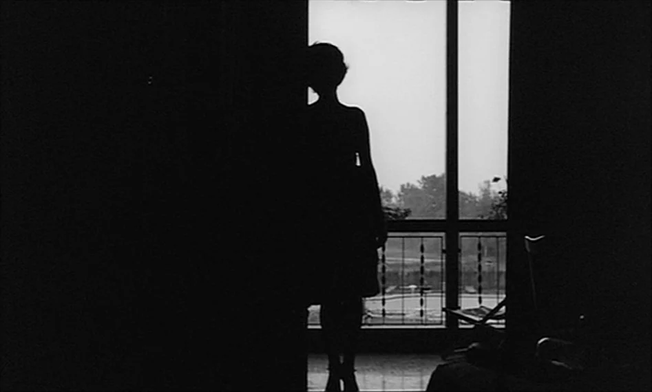 La Notte (1961) movie still: fullbody — A dark indoor space faces a bright glass balcony door overlooking a distant landscape. A woman stands in…