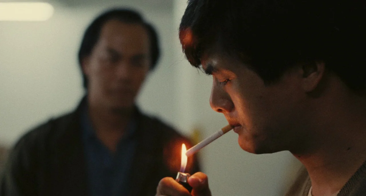 A Better Tomorrow (1986) movie still: closeup — A man holds a burning cigarette lighter to his cigarette inside a dark room. Another man stands in…