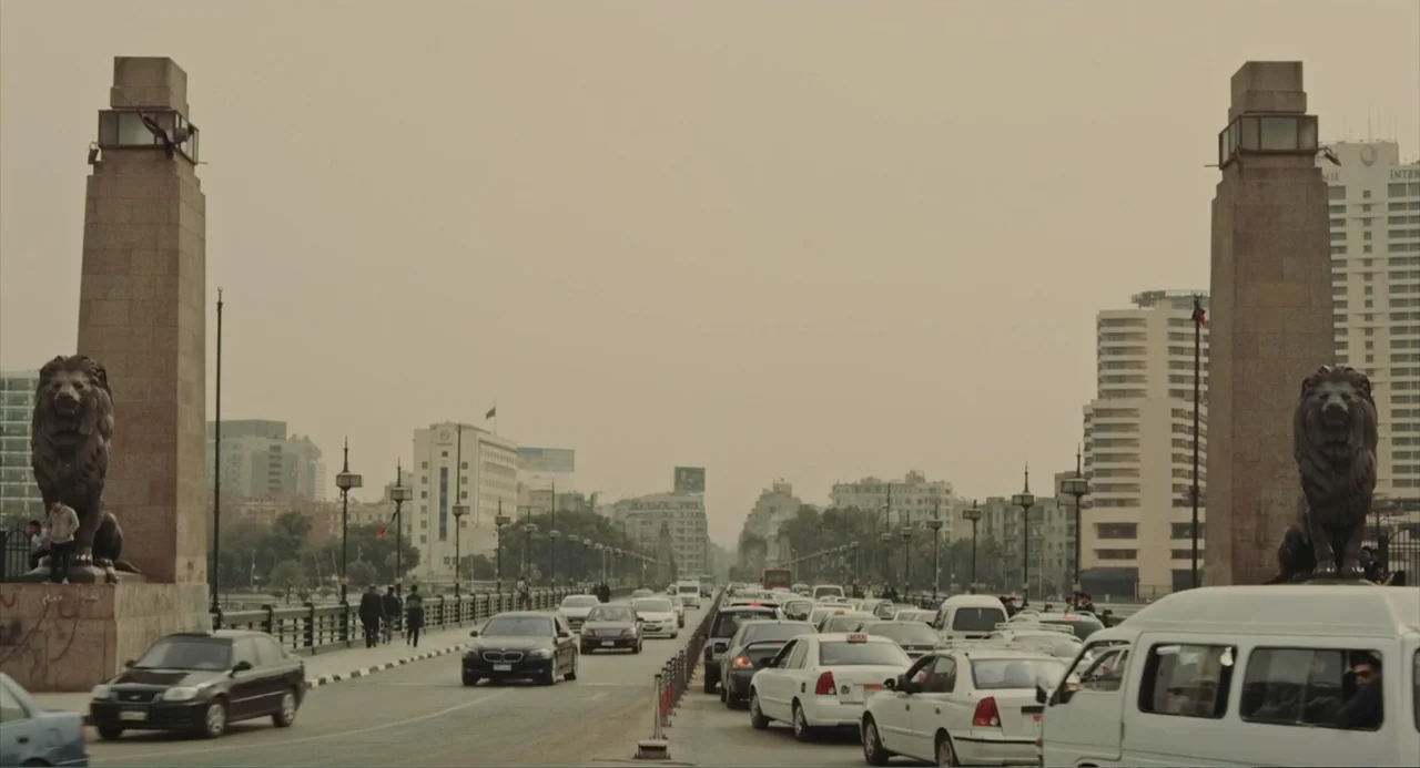 Cairo Jazzman (2017) movie still: wide — A wide shot shows a congested urban bridge with massive stone pedestals topped by lion statues. Numerous…