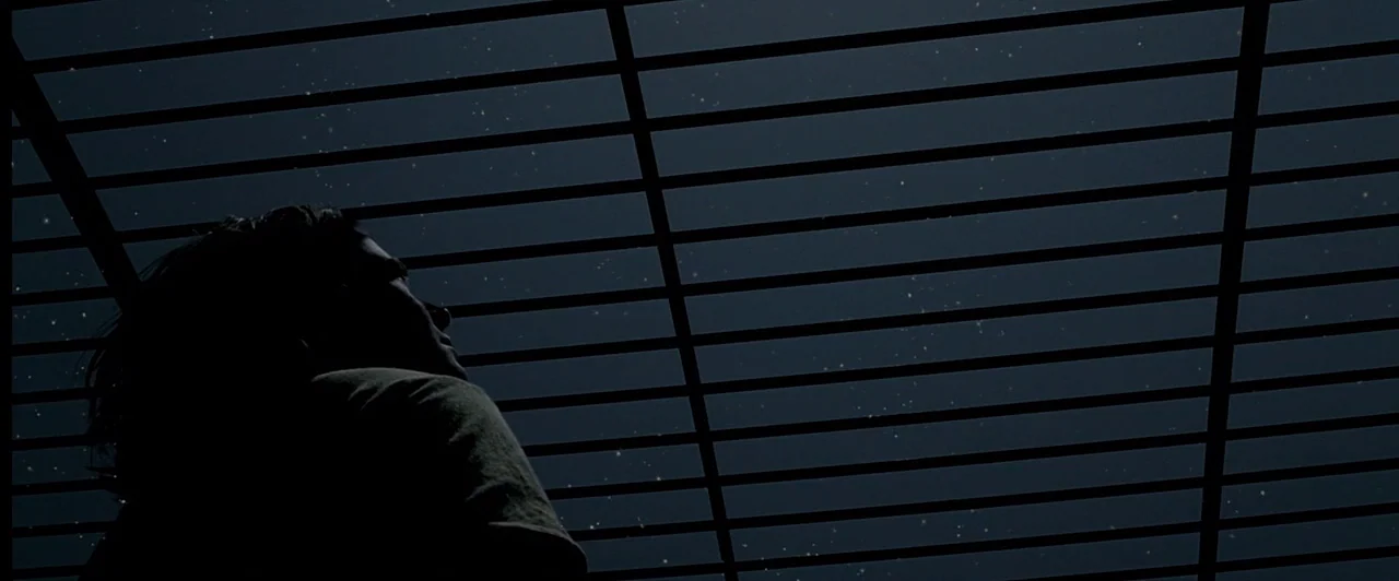 The Proposition (2005) movie still: medium — A night view of a dark sky seen through a horizontal metal grate structure. A person stands beneath the…
