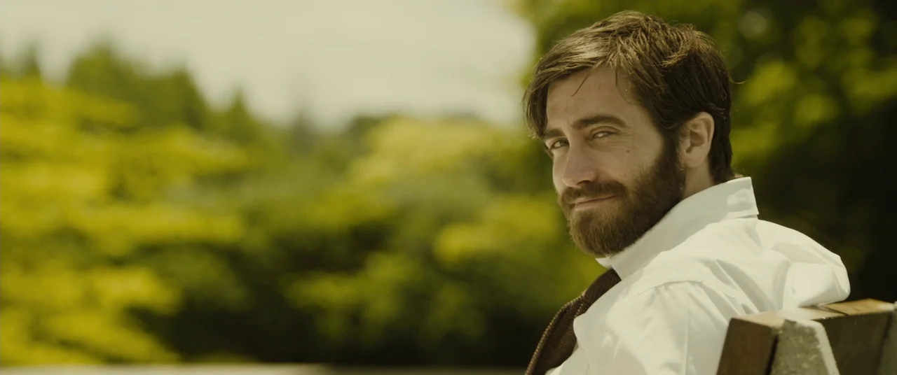 Enemy (2013) movie still: medium — A man sits on a wooden bench outdoors with blurred trees in the distance. The person turns his upper body to look…
