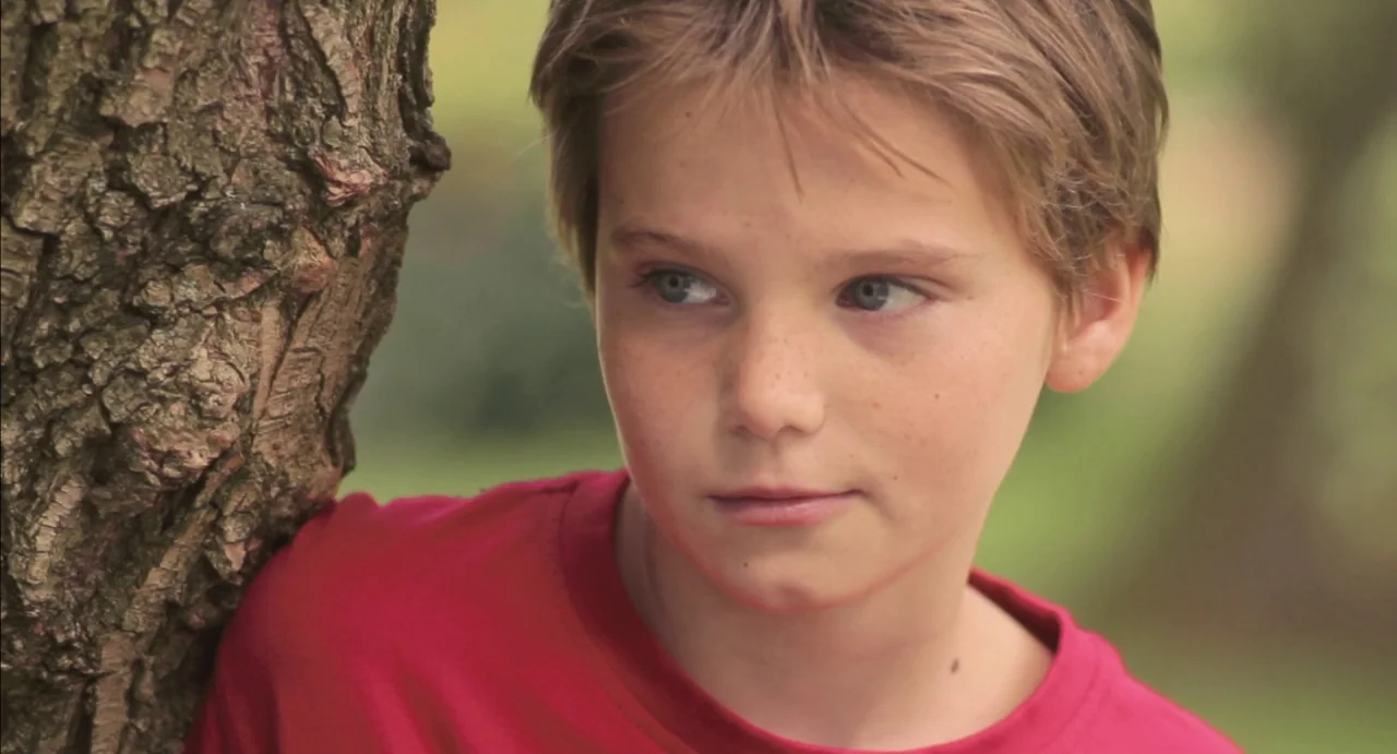 Tomboy (2011) movie still: closeup — A young boy stands next to a textured brown tree trunk in an outdoor setting. The child gazes toward the right…