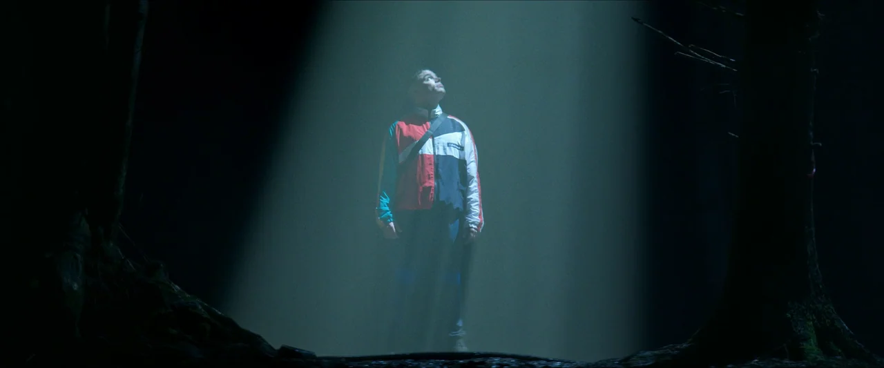 Kneecap (2024) movie still: fullbody — A dark, densely wooded area is lit by a single narrow overhead beam. A young man stands centered in the…