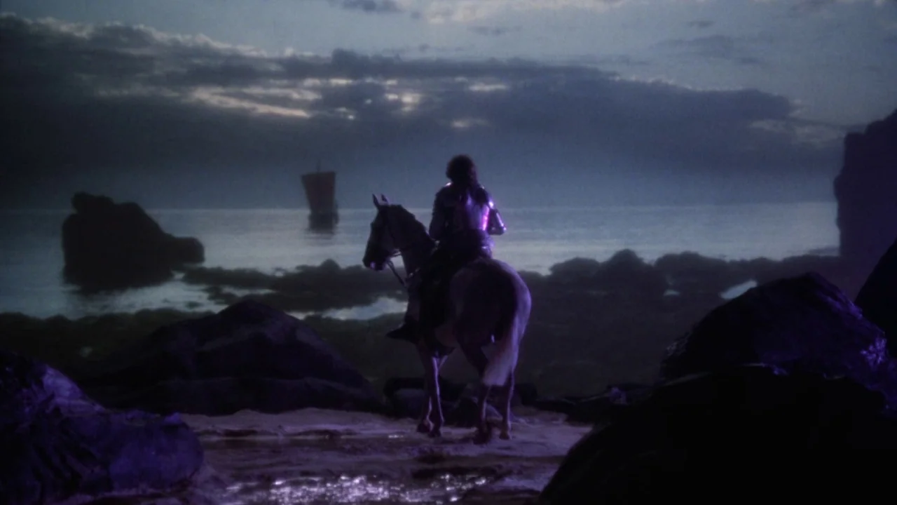 Excalibur (1981) movie still: wide — A rocky beach at dusk features a knight on horseback gazing at the water. In the distance, a small sailing ship…