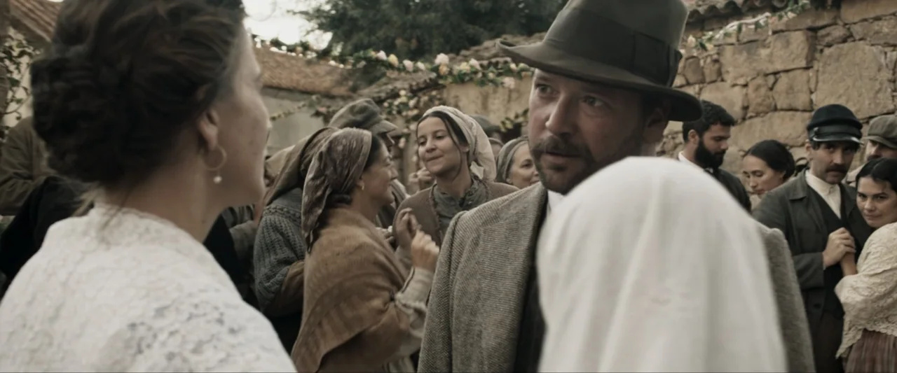Fatima (2020) movie still: medium — An outdoor village gathering with stone walls and floral decorations in the background. A man in a hat converses…