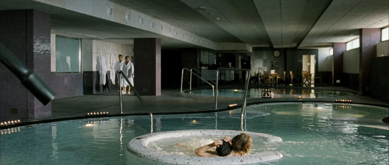Rivals (1972) movie still: wide — An indoor spa facility features multiple circular tiled pools surrounded by purple mosaic columns. A couple is…