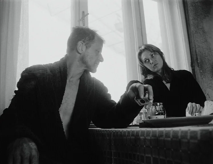 Damnation (1988) movie still: medium — Two people sit at a table covered by a checkered cloth in front of a window. The man reaches forward with a…