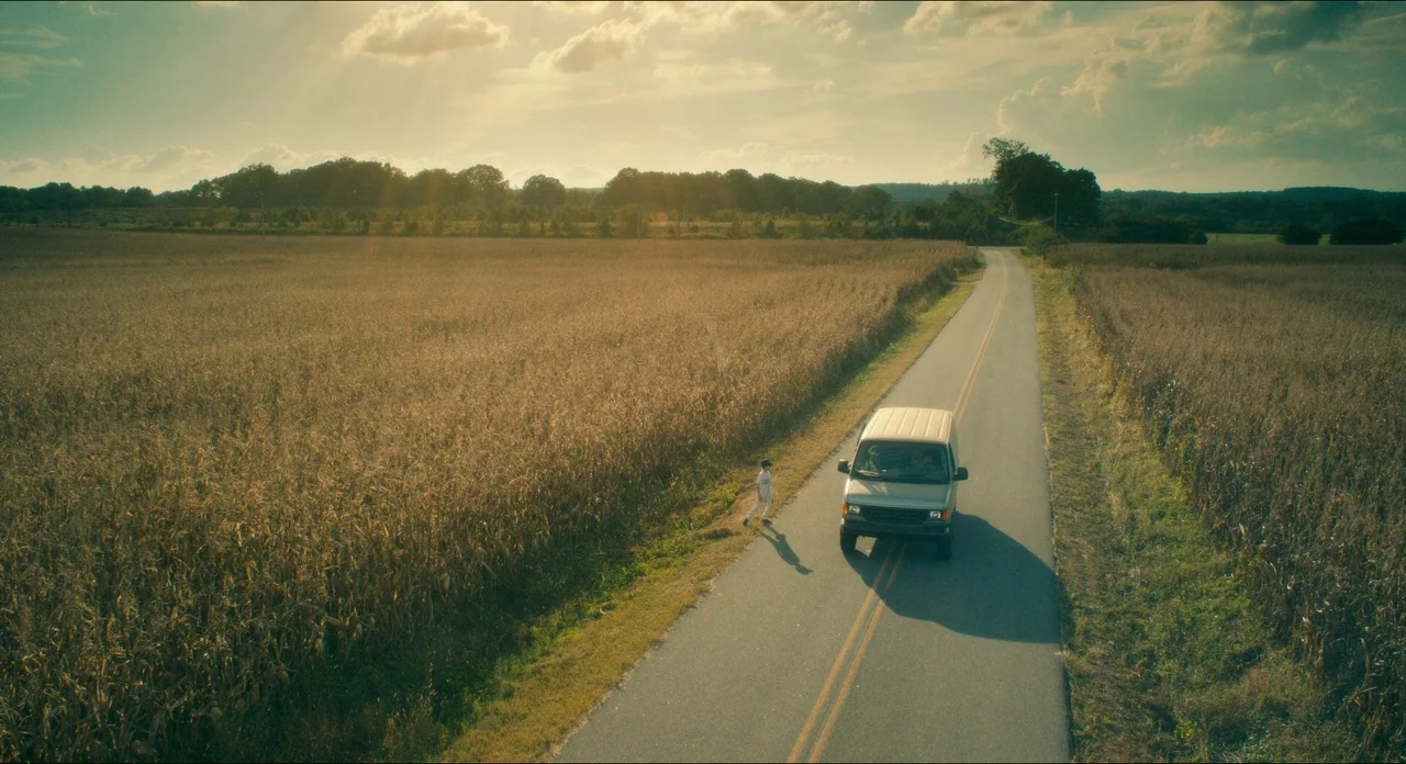 Doctor Sleep movie still: wide — A rural asphalt road cuts through a vast field of tall, dry cornstalks. A small child walks along the edge of the…