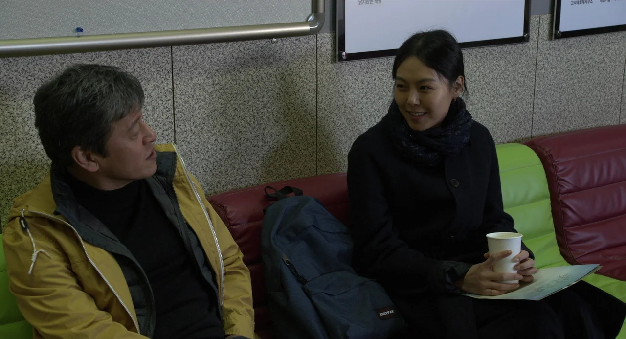 On The Beach At Night Alone (2017) movie still: medium — A waiting room with a textured grey wall and red and lime green seating. A man and a woman…