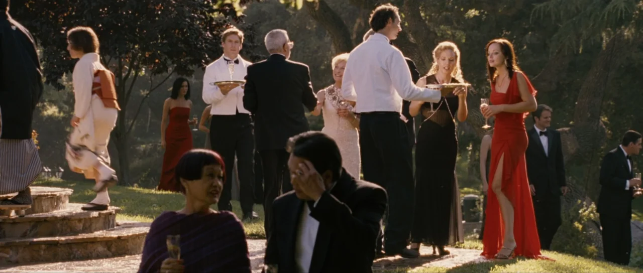 Monster-in-Law (2005) movie still: fullbody — A group of people in formal attire socialize on a sunny grassy lawn area. Guests hold glasses while…