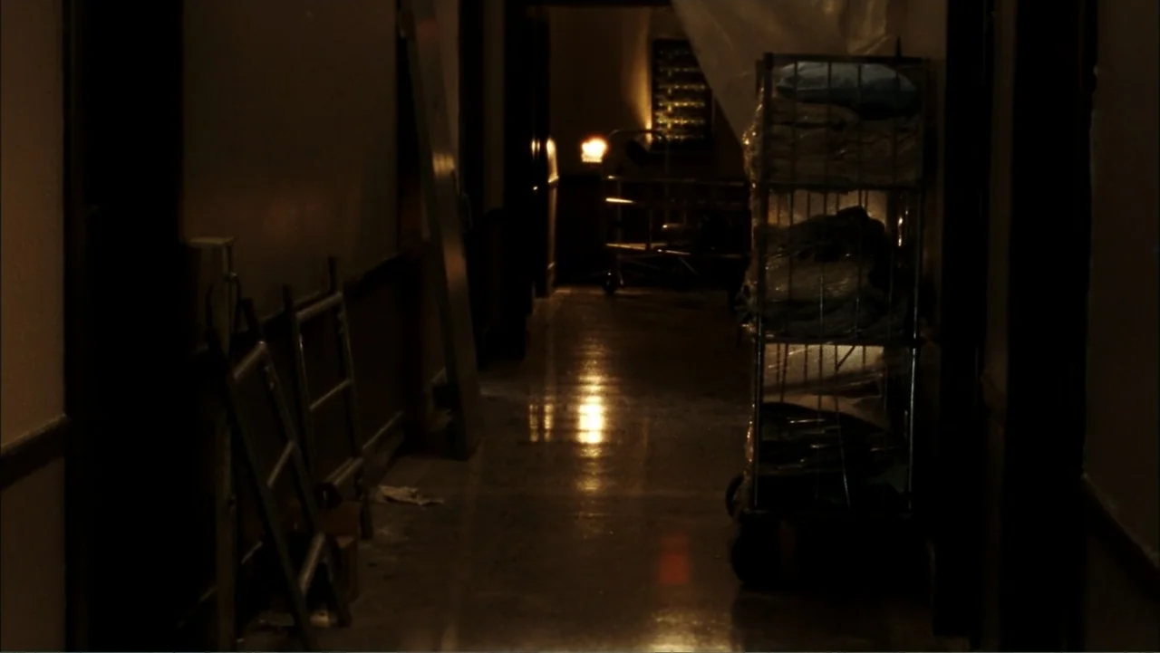 Driven to Kill (2009) movie still: establishing — A dimly lit, narrow hospital hallway features a metal linen cart on the right. An empty metal…
