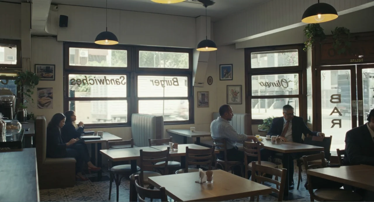 Belén (2025) movie still: wide — A quiet diner interior features multiple wooden tables and booths under pendant lights. Several customers sit…