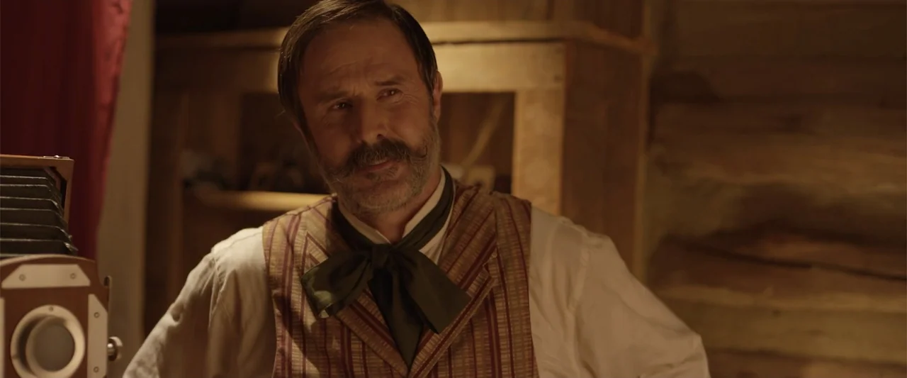 Ghosts of the Ozarks (2022) movie still: medium — A dimly lit room with wooden wall panels features a vintage bellows camera. A man dressed in…