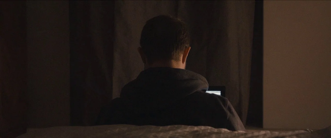 Simon Killer movie still: medium — A dark space features a person sitting on a bed facing a wall. The individual looks at the glowing screen of a…