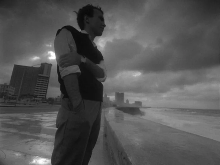 I Am Cuba (1964) movie still: medium — A seaside concrete walkway borders a turbulent ocean under heavy, overcast storm clouds. A young man wearing…