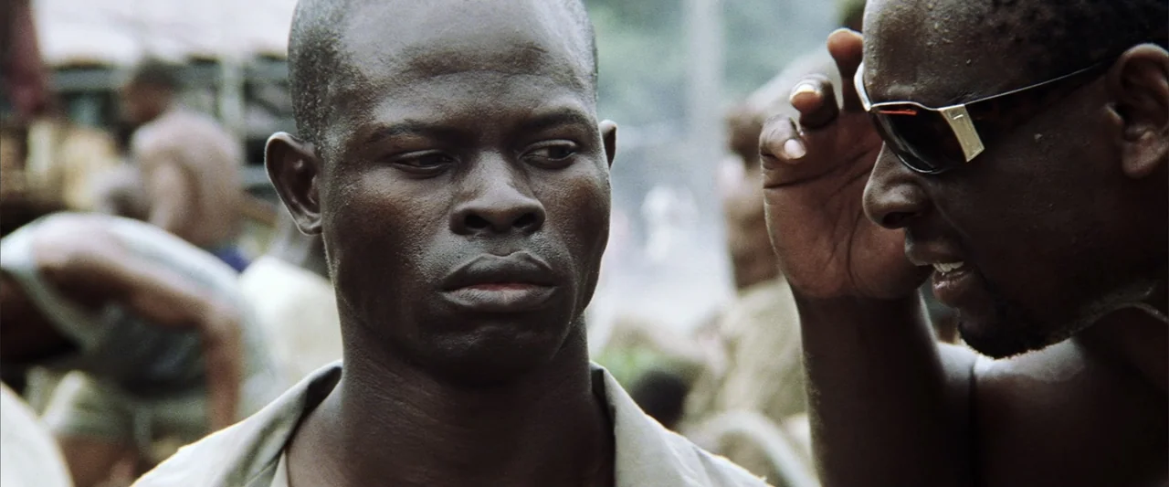 Blood Diamond (2006) movie still: medium — A crowded outdoor market setting with blurred people visible in the background. Two men engage in…